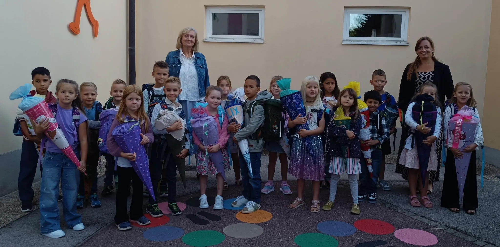 Eine Gruppe von Kindern und zwei Frauen stehen vor einem Gebäude. Die Kinder halten Blumensträuße und einige tragen Rucksäcke. Die Frauen tragen Jacken und eine hält ein Baby.