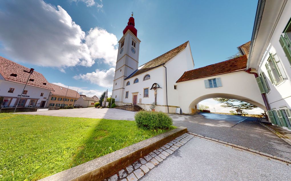 Abschnittsbild: Eine weiße Kirche mit einem roten Turm und Uhren, unter einem blauen Himmel mit Wolken, umgeben von einem grünen Rasen und einer gepflasterten Straße.