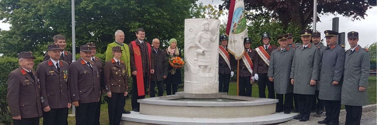 Eine Gruppe von Menschen in Uniformen und Mützen steht um ein Denkmal mit einer Skulptur eines Mannes, hält Blumen und Flaggen, in einem Park mit Bäumen im Hintergrund.