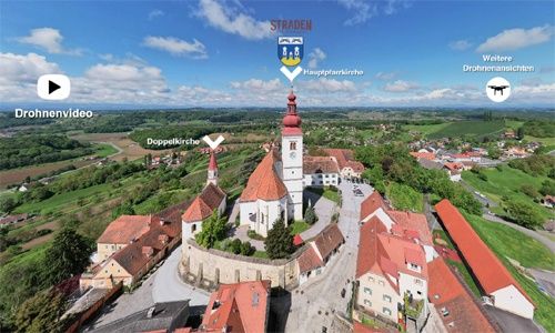 Eine Luftaufnahme des Dorfes Straden, Österreich, mit einer Kirche mit einem roten Turm, umgeben von rot gedeckten Gebäuden und üppiger Grünfläche. Das Bild enthält Beschriftungen für 'Drohnenvideo' und 'Doppelkirche'.