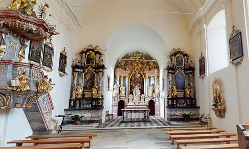 Ein prächtiger Innenraum einer Kirche mit goldenen Altären, Statuen und Gemälden an den Wänden. Der Altar hat einen goldenen Rahmen mit komplizierten Designs. Die Kirche hat Holzbänke und einen gemusterten Boden.
