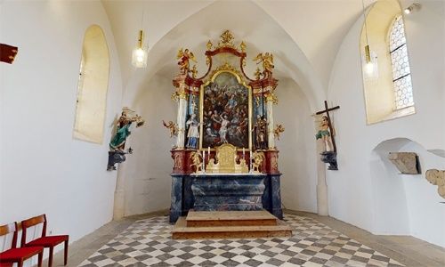 Ein kunstvoller Altar mit einem großen Gemälde und dekorativen Elementen steht in einer kleinen Kapelle. Die Kapelle hat einen gemusterten Boden und weiße Wände.