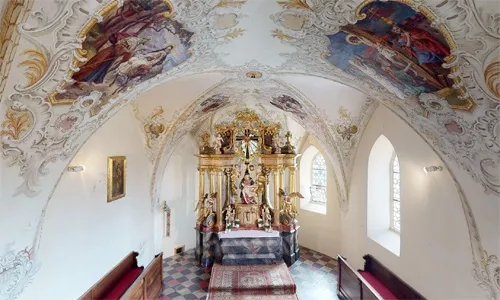 Ein kunstvoll verzierter Kapelleninnenraum mit einem goldenen Altaraufsatz, Fresken an der Decke und einem gemusterten Boden. Holzbänke sind vor dem Altar aufgestellt.
