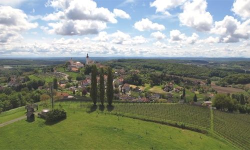 Luftaufnahme einer kleinen Stadt, umgeben von üppigen grünen Feldern und Weinbergen unter einem teilweise bewölkten Himmel.