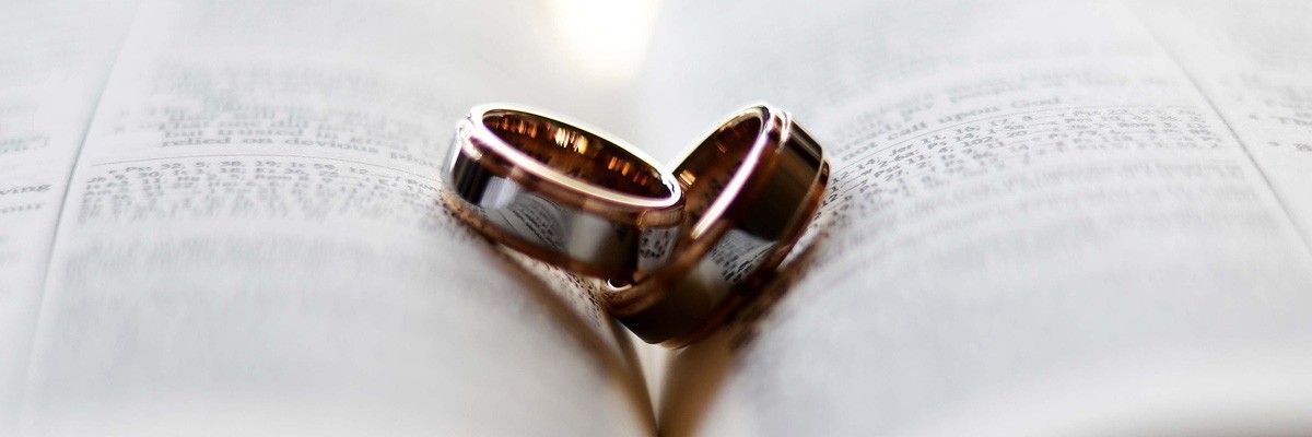 Zwei goldene Eheringe sind auf einem offenen Buch, möglicherweise eine Bibel, miteinander verflochten, wobei Text auf den Seiten sichtbar ist.