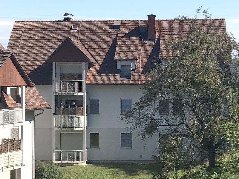 Ein großes weißes Mehrfamilienhaus mit braunen Dachziegeln und Balkonen, umgeben von Bäumen und Gras.