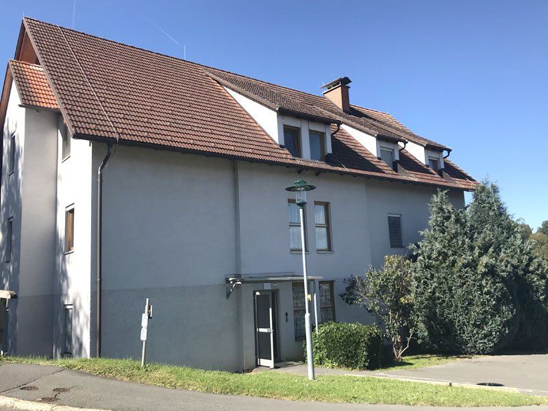 Ein modernes weißes Gebäude mit einem braunen Dach und mehreren Fenstern, an einem sonnigen Tag mit einer Straßenlaterne davor und Grünpflanzen drumherum.