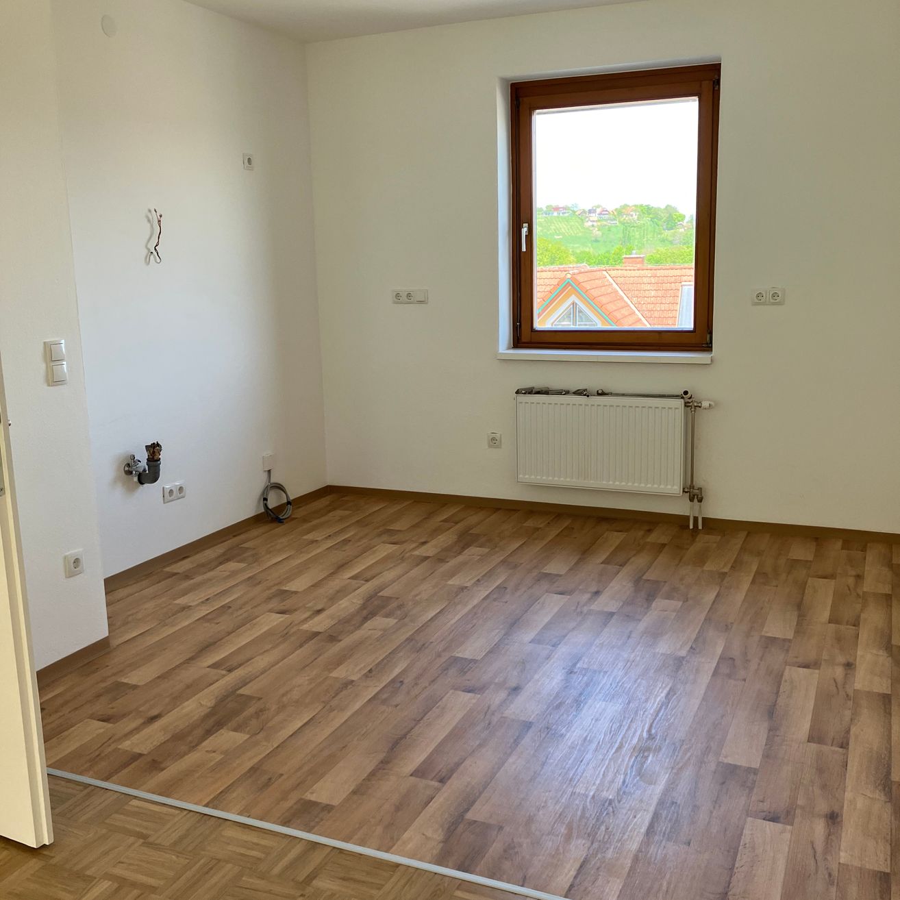 Ein leerer Raum mit Holzböden und weißen Wänden. Ein Fenster mit Blick auf ein Haus und Bäume befindet sich rechts. Ein Heizkörper ist unter dem Fenster.