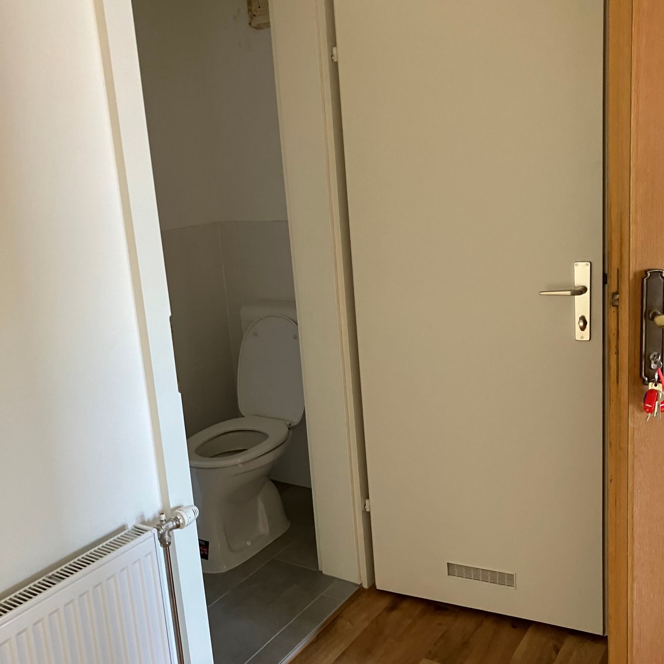 Ein kleines Badezimmer mit einer weißen Toilette und einem weißen Heizkörper auf der linken Seite. Die Badezimmertür ist offen und zeigt einen Holzboden.