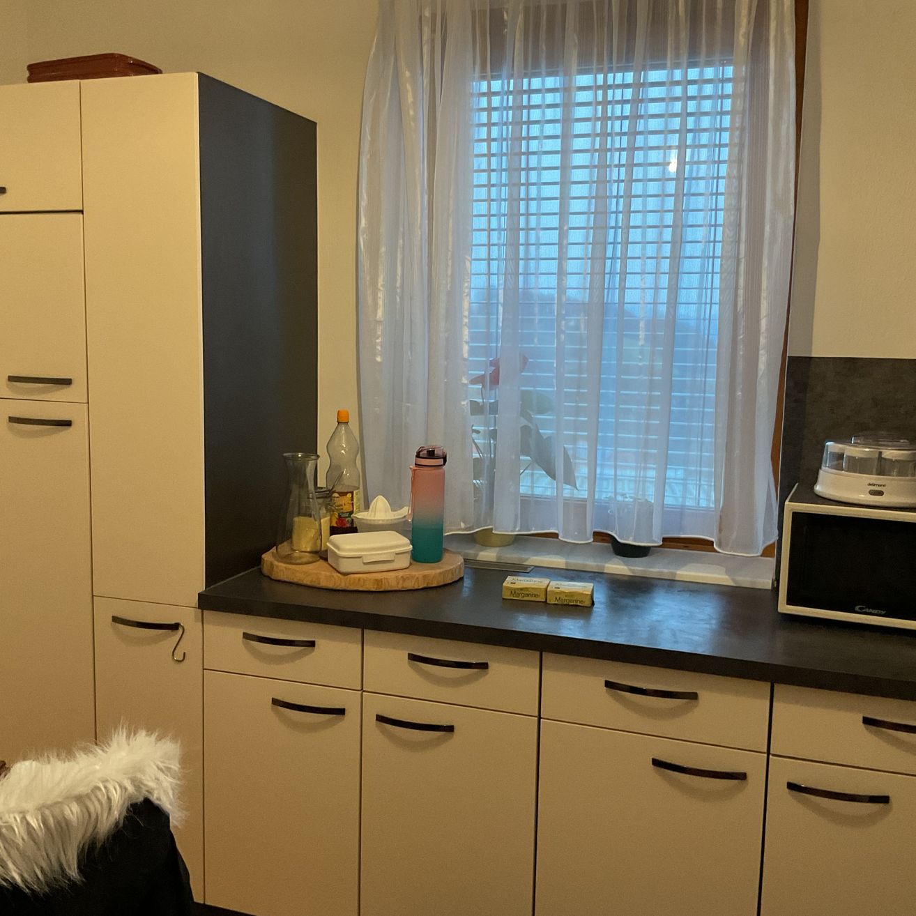 Eine Küche mit weißen Schränken, einer schwarzen Arbeitsplatte und einem Fenster mit weißen Vorhängen. Verschiedene Gegenstände stehen auf der Arbeitsplatte, darunter eine Flasche, ein Glas und eine Mikrowelle.