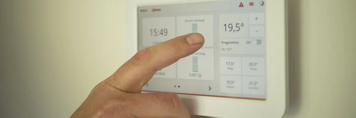 Eine Hand drückt einen Knopf auf einem digitalen Thermostaten, der die Zeit als 15:49 und die Temperatur als 19,5 Grad Celsius anzeigt.