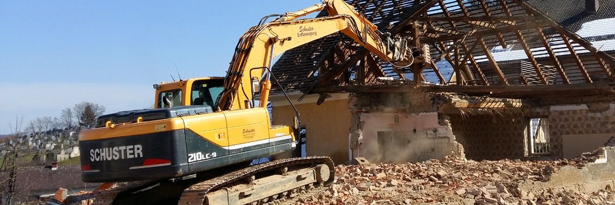 Ein gelber Bagger mit der Nummer 210LC-9 bricht ein Haus an einem sonnigen Tag ab. Der Bagger steht auf einem Schutthaufen, und das Haus wird gerade abgerissen.
