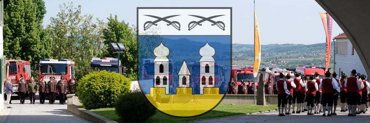 Ein Gemeindewappen mit zwei Schwertern und zwei Türmen wird auf einem Rasenbereich mit Menschen in traditioneller Kleidung und Feuerwehrfahrzeugen im Hintergrund gezeigt.