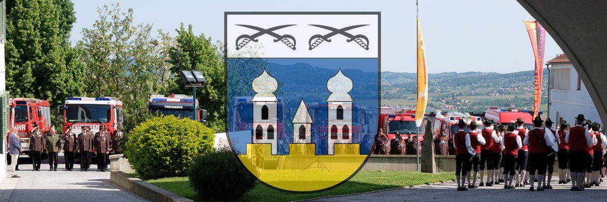 Ein Gemeindewappen mit zwei Schwertern und zwei Türmen wird auf einem Rasenbereich mit Menschen in traditioneller Kleidung und Feuerwehrfahrzeugen im Hintergrund gezeigt.