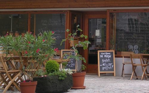 Ein Restaurant mit einer Tafel mit Speisekarte draußen, die verschiedene Gerichte anbietet, umgeben von Holztischen und Stühlen und Topfpflanzen.