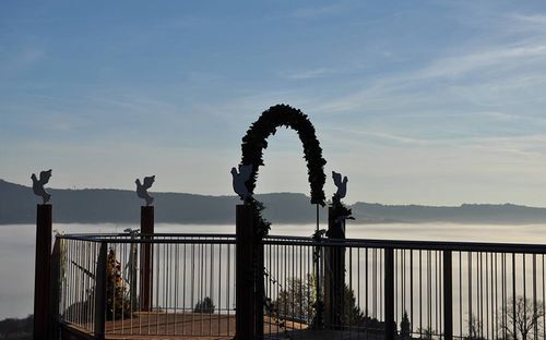 Eine Holzterrasse mit einem Metallgeländer und einem dekorativen Bogen, geschmückt mit Grünpflanzen und Engelsfiguren. Die Terrasse bietet einen Blick auf eine neblige Landschaft mit Bergen in der Ferne.