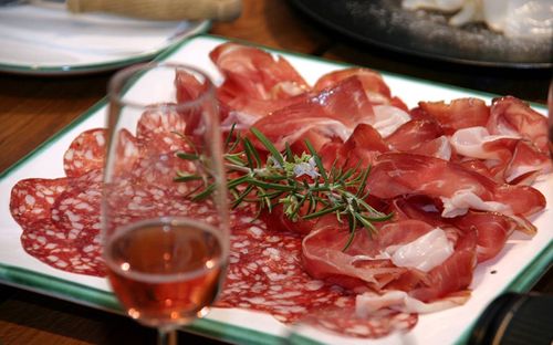 Ein Teller mit geschnittenen Fleischsorten, einschließlich Prosciutto und Salami, garniert mit einem Rosmarinzweig, begleitet von einem Glas Wein auf einem Holztisch.