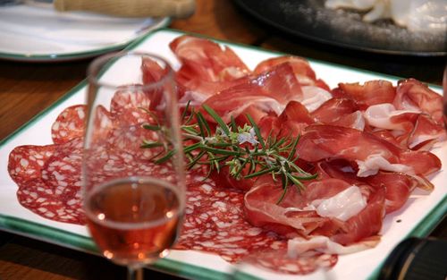 Ein Teller mit geschnittenen Fleischsorten, einschließlich Prosciutto und Salami, garniert mit einem Rosmarinzweig, begleitet von einem Glas Wein auf einem Holztisch.