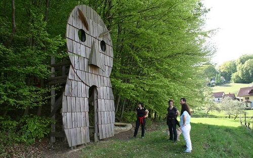 Vier Personen stehen vor einer Holzskulptur eines Gesichts im Wald, eine Frau hält einen Hund.