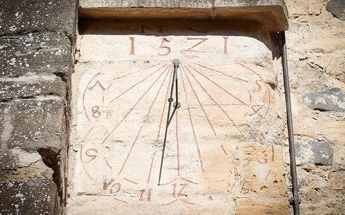 Eine alte Sonnenuhr an einer Steinmauer, mit Zahlen von 1 bis 12 und einem zentralen Gnomon, der einen Schatten wirft.