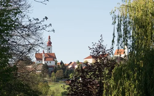 Ein malerisches Dorf mit einer Kirche und roten Dächern, umgeben von üppigem Grün unter einem klaren blauen Himmel.