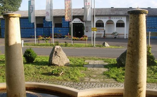 Ein Museumseingang mit zwei großen Säulen, einem Brunnen und einem großen Felsen im Vordergrund. Das Gebäude hat eine blaue Wand und ein Schild, das 'Johannisbrunnen Museum' besagt.