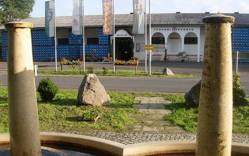 Ein Museumseingang mit zwei großen Säulen, einem Brunnen und einem großen Felsen im Vordergrund. Das Gebäude hat eine blaue Wand und ein Schild, das 'Johannisbrunnen Museum' besagt.