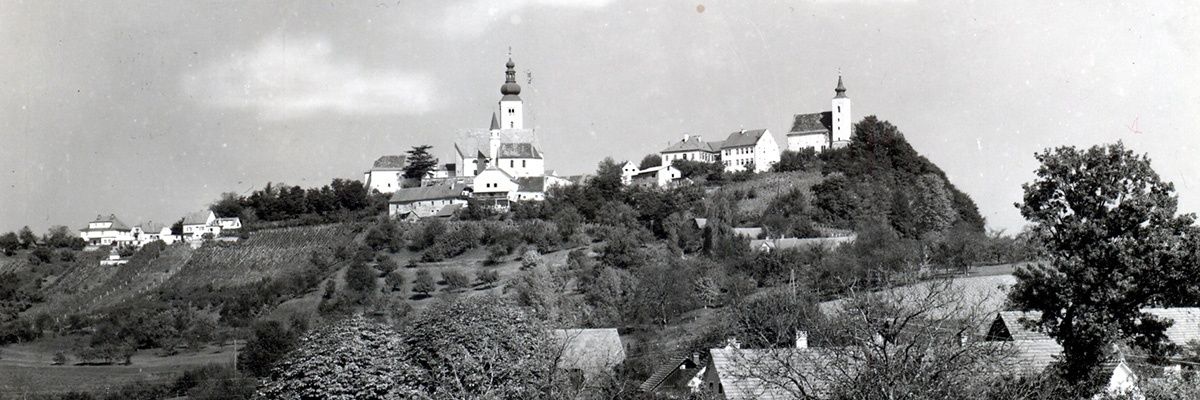 Ein Schwarz-Weiß-Foto zeigt ein Dorf auf einem Hügel mit mehreren Gebäuden, darunter eine Kirche mit einem Turm, umgeben von Bäumen und Grün.