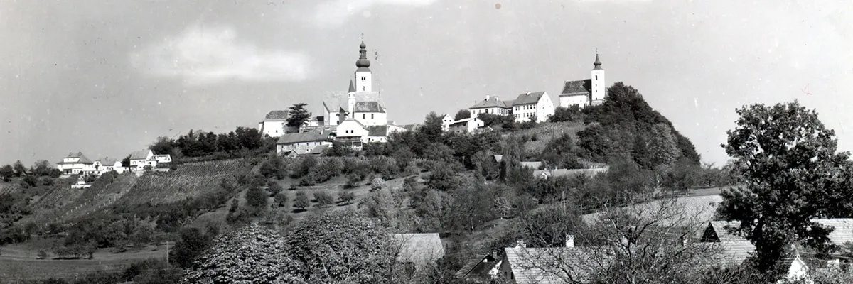 Ein Schwarz-Weiß-Foto zeigt ein Dorf auf einem Hügel mit mehreren Gebäuden, darunter eine Kirche mit einem Turm, umgeben von Bäumen und Grün.