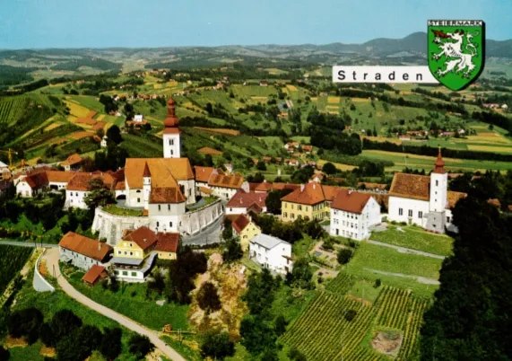 Eine Luftaufnahme des Dorfes Straden mit einer Kirche, Häusern und Weinbergen vor einer Kulisse aus grünen Hügeln und blauem Himmel.