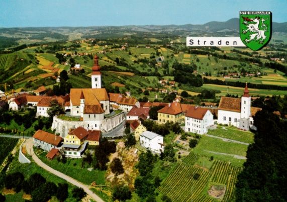 Eine Luftaufnahme des Dorfes Straden mit einer Kirche, Häusern und Weinbergen vor einer Kulisse aus grünen Hügeln und blauem Himmel.