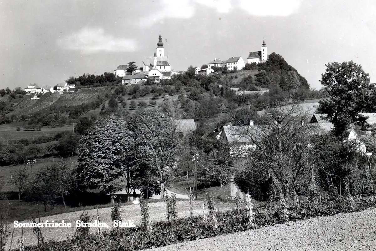 Ein Schwarz-Weiß-Foto eines Dorfes auf einem Hügel mit einer Kirche in der Mitte, umgeben von Bäumen und Häusern.