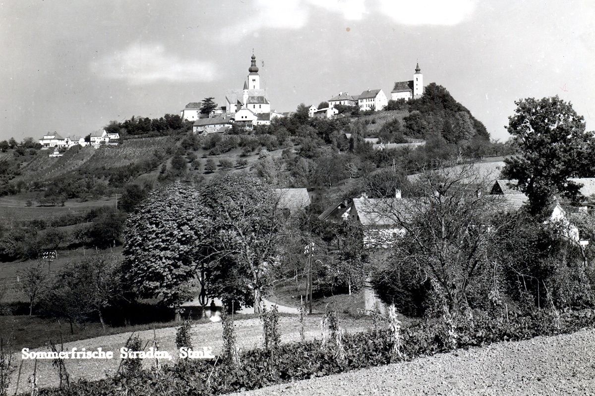 Ein Schwarz-Weiß-Foto eines Dorfes auf einem Hügel mit einer Kirche in der Mitte, umgeben von Bäumen und Häusern.