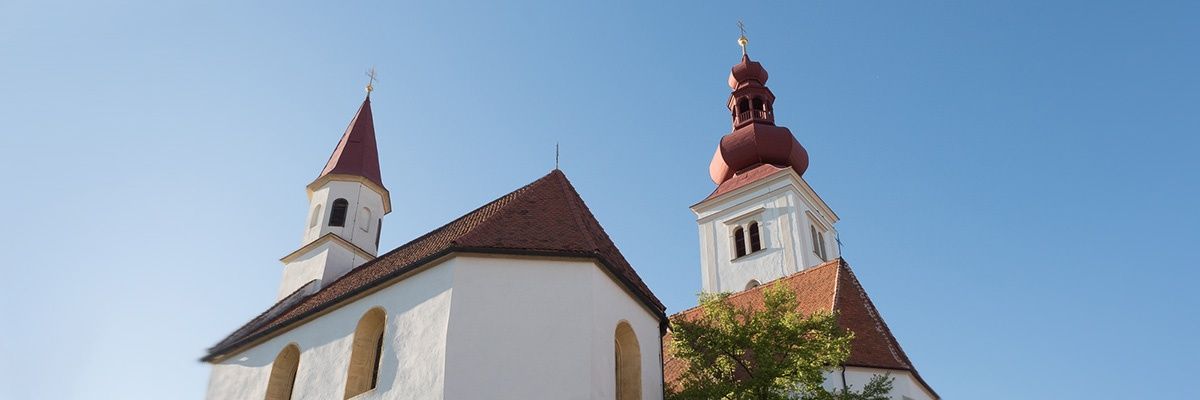 Eine Kirche mit zwei Türmen, einer mit einer roten Kuppel und der andere mit einem spitzen Dach. Die Kirche hat ein braunes Dach und weiße Wände mit Bogenfenstern.