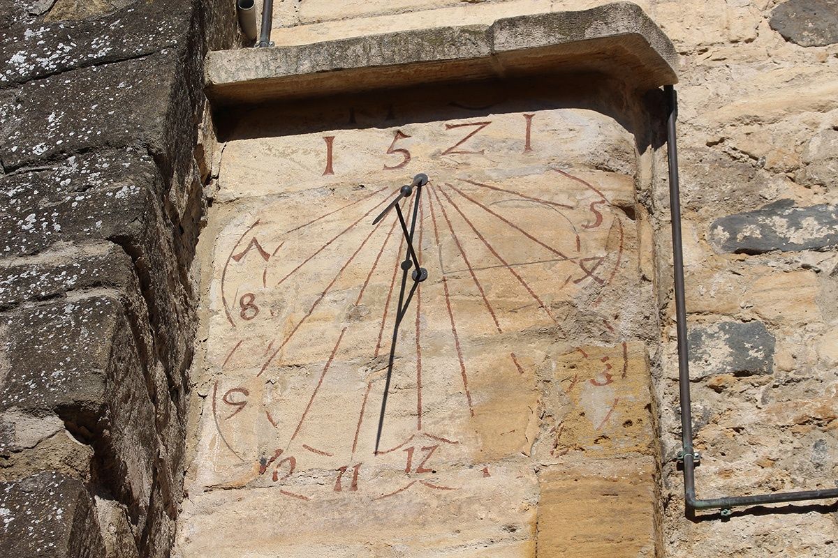 Ein verwittertes Sonnenuhr mit römischen Ziffern von 1 bis 12, an einer Steinmauer montiert. Der Gnomon zeigt auf die Zahl 11.