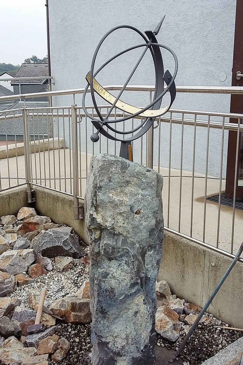 Eine Sonnenuhr mit einem goldenen Ring ist auf einem großen grauen Stein montiert, umgeben von Steinen und einem Metallgeländer.