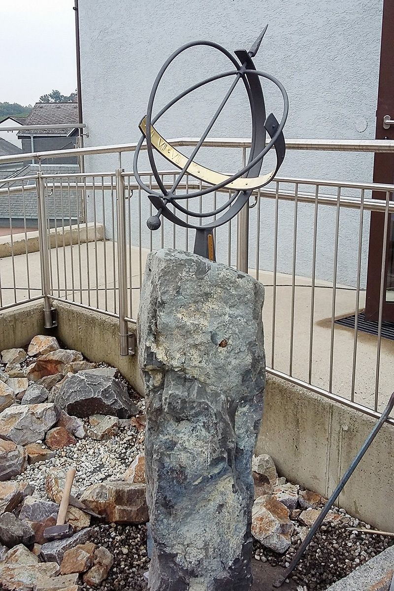 Eine Sonnenuhr mit einem goldenen Ring ist auf einem großen grauen Stein montiert, umgeben von Steinen und einem Metallgeländer.