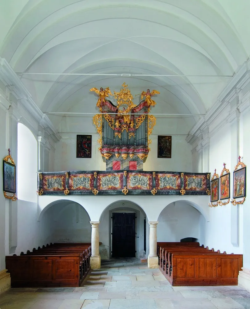 Innenansicht einer Kirche mit einer großen, verzierten Orgel über dem Eingang, umgeben von gewölbten hölzernen Kirchenbänken und geschmückt mit gerahmten Gemälden an den Wänden.