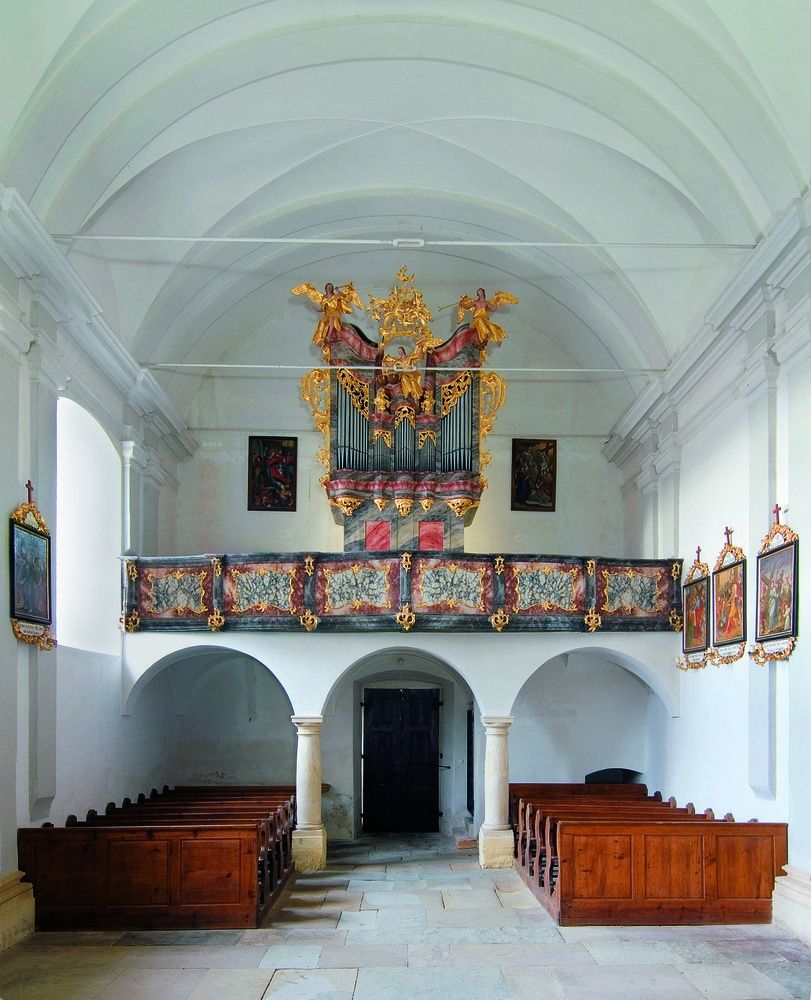 Innenansicht einer Kirche mit einer großen, verzierten Orgel über dem Eingang, umgeben von gewölbten hölzernen Kirchenbänken und geschmückt mit gerahmten Gemälden an den Wänden.