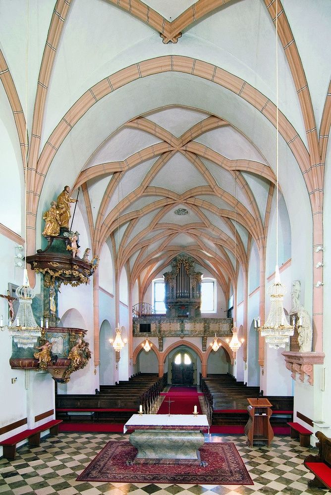 Das Innere einer Kirche mit hoher Decke, Holzbögen und einem roten Teppich, der zum Altar führt. Es gibt Statuen, Kronleuchter und eine Orgel.