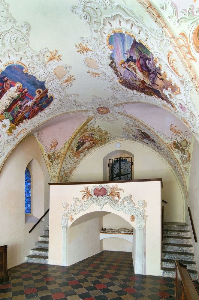 Eine kunstvoll verzierte Kapellendecke mit aufwendigen Fresken, die religiöse Szenen darstellen, einem zentralen Bogen und einer Treppe, die zu einem kleinen Altar führt.