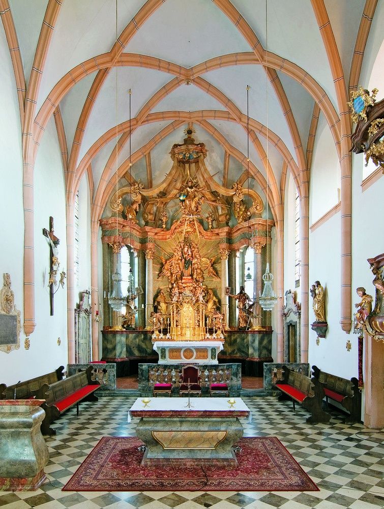Ein prächtiger Kircheninnenraum mit goldenem Altarbild, kunstvollen Holzschnitzereien und Statuen. Der Altar ist von einem roten Teppich und einem weißen Tuch umgeben.