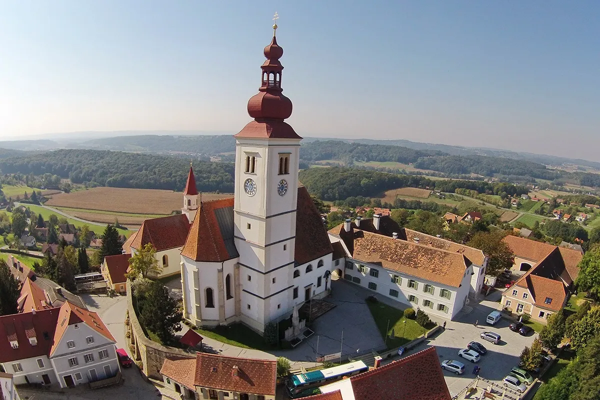 Ein Luftbild einer historischen Kirche mit einem hohen Turm, vor dem Hintergrund saftiger grüner Hügel und eines klaren blauen Himmels.