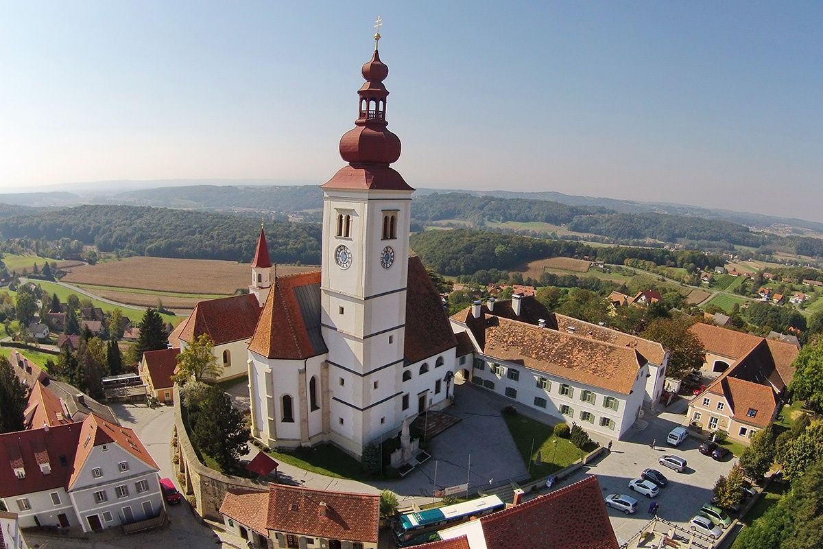 Ein Luftbild einer historischen Kirche mit einem hohen Turm, vor dem Hintergrund saftiger grüner Hügel und eines klaren blauen Himmels.