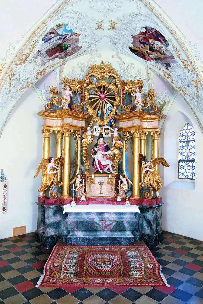 Ein prächtiger goldener Altar in einer Kirche mit einer zentralen Statue einer Frau, die ein Kind hält, umgeben von Engeln und Cherubs, mit kunstvollen Schnitzereien und einem Buntglasfenster im Hintergrund.