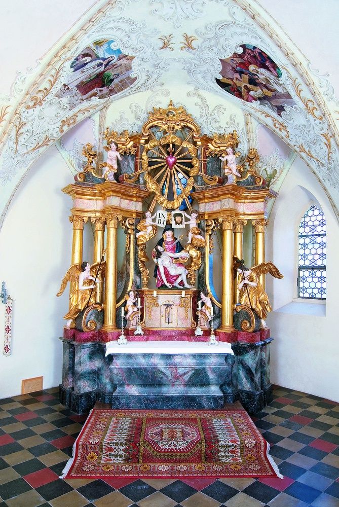 Ein prächtiger goldener Altar in einer Kirche mit einer zentralen Statue einer Frau, die ein Kind hält, umgeben von Engeln und Cherubs, mit kunstvollen Schnitzereien und einem Buntglasfenster im Hintergrund.