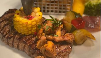 Ein Nahaufnahme eines perfekt gegrillten Steaks, das mit Mais, Pilzen und Gemüse auf einem weißen Teller belegt ist.