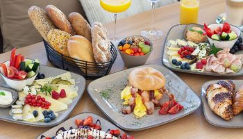 Ein Holztisch mit einer Auswahl an Frühstücksgerichten, darunter Brot, Obst, Eier und Gebäck. Es gibt auch Gläser Orangensaft und Wein.