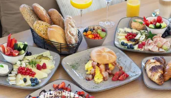 Ein Holztisch mit einer Auswahl an Frühstücksgerichten, darunter Brot, Obst, Eier und Gebäck. Es gibt auch Gläser Orangensaft und Wein.