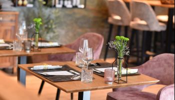Ein Restaurant mit Holztischen und Stühlen, mit einer Vase mit Blumen, Weingläsern und Besteck auf dem Tisch. Die Stühle sind rosa und die Wände sind aus Stein.